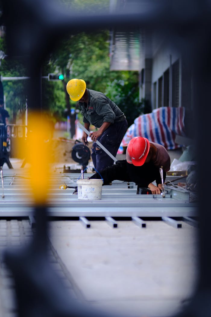Two construction workers in helmets working on an outdoor site with tools and materials visible.